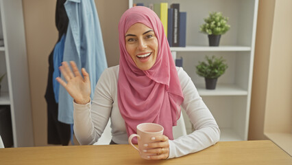 Wall Mural - Smiling woman wearing hijab greeting while enjoying coffee at home, evoking warmth and friendliness.