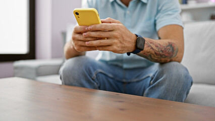 Wall Mural - A tattooed young man uses a smartphone in a modern living room, with a subtle beard and casual attire reflecting a relaxed home setting.