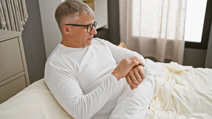 Sticker - Mature grey-haired man in white shirt relaxing on bed in a well-lit bedroom interior