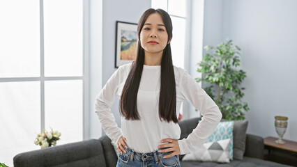 Canvas Print - Confident asian woman standing in a modern living room with hands on hips and natural light flooding in