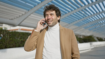 Wall Mural - Smiling hispanic man in casual fashion talking on phone outdoors in a city park.