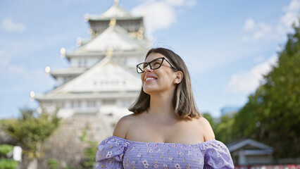 Poster - Confident and joyful hispanic woman in glasses, smiling while posing happily at famous osaka castle, her carefree expression reflecting the beauty of japan's heritage.