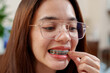© Ekkasit A Siam - Asian girl with glasses closely inspecting braces in small mirror, using toothpick. Focus on dental care and personal hygiene routine. Casual home setting with neutral background.