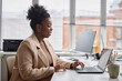 © Seventyfour - Modern Office: Side view of young Black businesswoman sitting at laptop. Smiling successful manager typing emails
