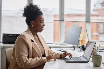 Poster - Modern Office: Side view of young Black businesswoman sitting at laptop. Smiling successful manager typing emails