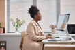 © Seventyfour - Side view of adults African American woman in beige fashionable suit sitting at working desk in front of computer screen in office typing on keyboard and networking, copy space