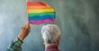 © Anastasia Knyazeva - Older adult proudly displays a rainbow flag against a backdrop of a clear blue sky. Their joyful smile, stylish sunglasses, and warm scarf highlight their vibrant personality and the essence of LGBTQ+