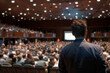 © Babb - A man stands in front of a large crowd of people, giving a presentation