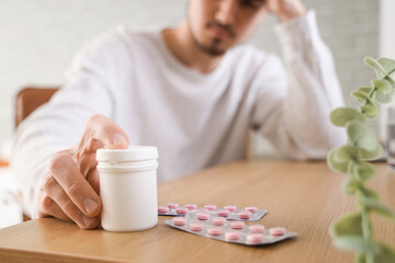 Poster - Young man with headache reaching for pills at home, closeup