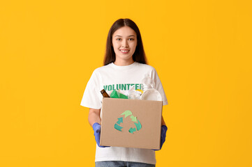 Young female volunteer holding recycle box with garbage on yellow background