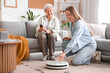 © Pixel-Shot - Young woman showing her grandmother how to use robot vacuum cleaner at home