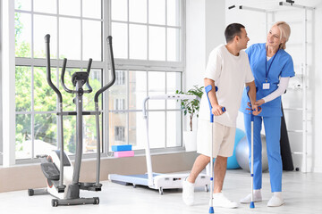 Wall Mural - Female physiotherapist and young man with crutches walking in rehabilitation center