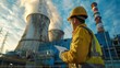 © mongkonchai - Nuclear power plant engineer A man with a beard in a protective helmet and an orange vest stands against the background of a nuclear power plant. Work inspection