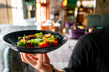 Wall Mural - Close-up of a hand holding a black plate with a gourmet dish featuring grilled meat and fresh vegetables, set against a blurred restaurant background