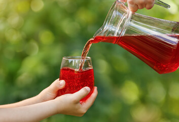 Wall Mural - child hands holding glass and cherry juice pouring from jug with green garden on the background