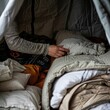 © Parintron - Person arranging sleeping area in a cozy tent with pillows and blankets, preparing for a comfortable night in a camping adventure.