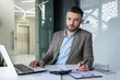 © Liubomir - Confident businessman working in a modern office. He is using a laptop and reviewing documents at the desk.