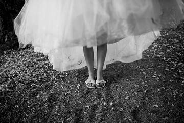 Wall Mural - A bride stands on a path covered with leaves, lifting her wedding dress slightly to reveal bright pink sandals underneath, adding a touch of playful personality to her elegant attire.