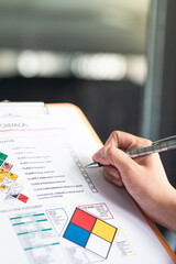 Action of a safety supervisor hand is checking on checklist of chemical and hazardous material form to verify the risk with blurred background of factory storage workplace. Industrial working scene.