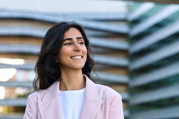 Wall Mural - Successful smiling indian or arabic woman in casual business suit looking aside. Closeup headshot outdoor portrait of young middle eastern Israel businesswoman standing office building. Copy space