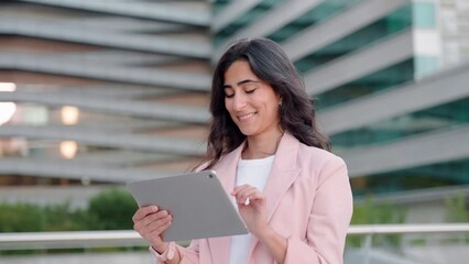 Wall Mural - Smiling young middle eastern Israel businesswoman using tablet pc application for online remote work standing at office business building outdoors. Indian or arabic 30s woman holding digital computer