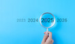 © Supatman - The 2025 New Year business goals concept. Hand holding a magnifying glass highlighting year 2025 with a target icon, representing focus on business goals and objectives for the future. Marketing trend