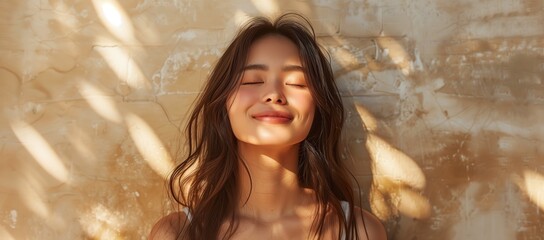 Wall Mural - A beautiful Asian woman with long hair smiles and looks at the sun with her eyes closed. She stands in front of a beige wall with sunlight falling on it.