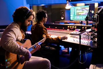 Wall Mural - Skilled artist musician recording his guitar sounds in professional studio, using electro acoustic tunes to edit another record for an album. Singer composing music with audio expert in control room.