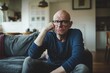 © pham - A middle-aged man with glasses sits on a couch in a living room, looking thoughtful and concerned