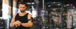 © Prostock-studio - A young man with a beard is wearing a black tank top and checking his smartwatch. He is standing in a gym, with exercise equipment blurred in the background, copy space