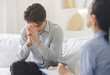 © Prostock-studio - A distressed man sits on a couch with his head in his hands, engaging in a conversation with a counselor who takes notes. The setting appears to be a bright