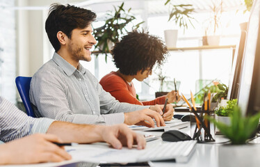 Aufkleber - Multiethnic men are seated at a desk, engrossed in their work on computers. Each person is focused on their screen, typing and clicking away, with papers and notebooks scattered around them.