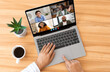 © Prostock-studio - Woman using laptop for a video chat with multiple participants. The laptop is open on a wooden desk with a cup of coffee and a potted plant nearby, top view