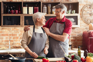 Wall Mural - A woman is chopping a cucumber on a wooden cutting board in a kitchen, while a man in the background prepares a salad. The kitchen is bright and sunny, and the scene is one of peaceful domesticity.