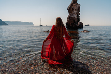 Wall Mural - A woman in a red dress stands on a beach with a rocky shoreline in the background. The scene is serene and peaceful, with the woman's red dress contrasting against the natural elements of the beach.