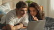 © Yuliia - Two people couple man woman looking at laptop screen girlfriend show finger discussing talking chatting watching movie film together weekend home bedroom lying on bed relax boyfriend male female smile