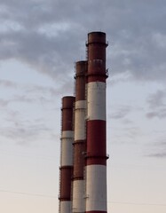 Three tall industrial chimneys stand against a cloudy sky, showcasing their weathered red and white bands. The overcast atmosphere adds a dramatic and somber mood to the scene.