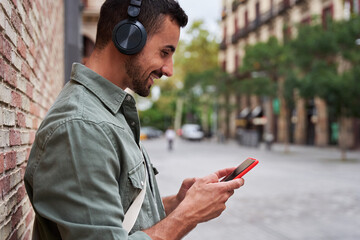 Cheerful caucasian man relaxing outdoors using phone sending text messages and checking the social media and listening music leaning in a brick wall in an urban street. Copy space, side view image.