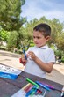 © Diego Schiochet - Boy with white polo concentrated in a craft activity on a wooden table in the park. She has blue scissors in her hand, ready to use, and colored crayons scattered on the table.