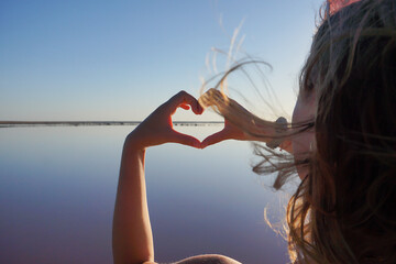 The girl holds her hands in the shape of a heart against the background of a pink lake.