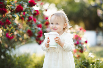 Poster - Smiling baby girl 2-3 year old holding cup of coffee outdoor over sunny nature background close up. Summer season. Childhood.