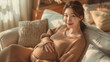 © Oriol Roca - Pregnant Asian woman sitting on her living room couch wraps her hands around her belly.