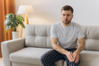 © anatoliycherkas - Unhappy man sitting on sofa at home and touching hurt knee with grimace of pain and suffering. Young guy with rheumatism, arthritis or osteoarthritis feeling intense severe pain in knee joint