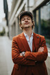 © qunica.com - Confident young businessman wearing an orange suit and cap, standing outdoors with arms crossed, looking self-assured and professional.
