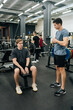 © dikushin - Full length vertical shot of muscular personal instructor explaining technique of exercise to young beginner sportsman sitting on bench with barbell during fitness training. Concept of healthy life.