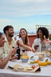 © CarlosBarquero - Vertical. Laughing group diverse young friends enjoying lunch together outdoors. Cheerful people gathered drinking red wine eating snack on summer day having fun celebrating birthday party on rooftop