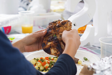 close-up of child's hands holding grilled chicken, background view of plate with corn salad, carrots and green beans