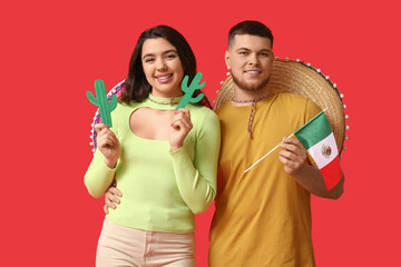 Canvas Print - Beautiful young happy couple with sombrero hats, paper cacti and Mexican flag on red background. Cinco de Mayo celebration