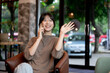© bongkarn - An attractive Asian woman is waving her hand to greet someone while talking on the phone in a cafe.