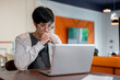 © bongkarn - A thoughtful young Asian man is working on his laptop, looking at the screen with a serious face.